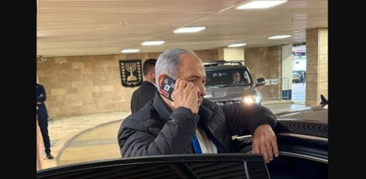φωτογραφία δείχνει τον Benjamin Netanyahu να μιλά σε κινητό με κόκκινη ταινία στις κάμερες.