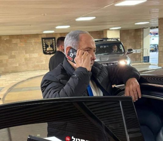 φωτογραφία δείχνει τον Benjamin Netanyahu να μιλά σε κινητό με κόκκινη ταινία στις κάμερες.